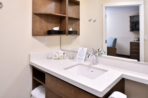 Un baño de hotel con un lavamanos blanco, un espejo, un estante de madera con artículos de aseo y un perchero para toallas.