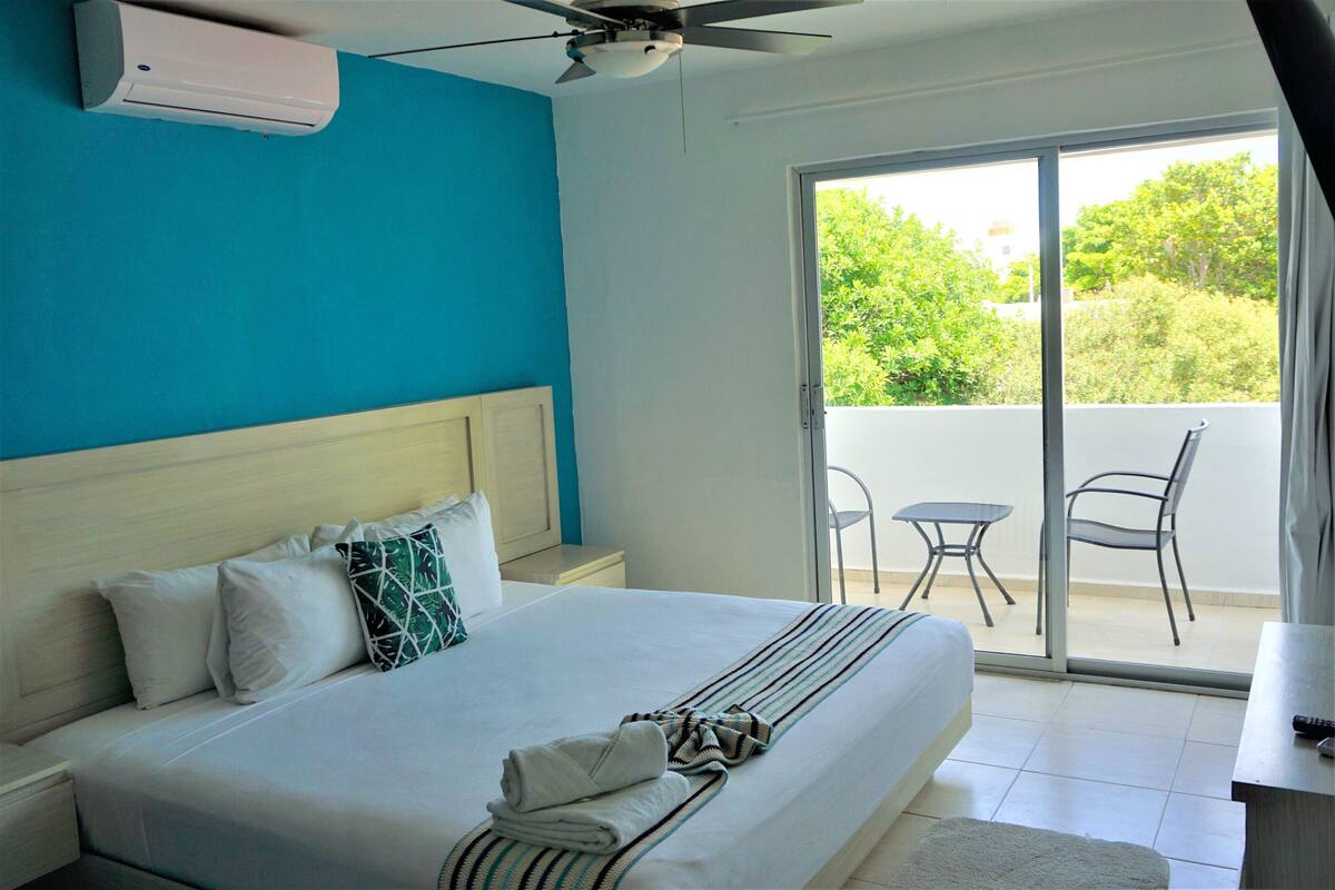 A hotel room with a bed, a ceiling fan, a window with a view of greenery, and a small table with a chair.