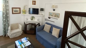 A living room with a sofa, armchair, and coffee table. There is a TV on a stand, a lamp, and framed pictures on the wall.