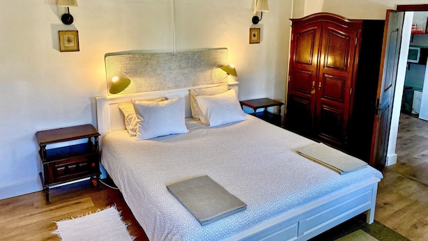 A neatly made bed with a patterned bedspread, two bedside tables with lamps, a wooden wardrobe, and a white door leading to a kitchen.