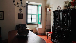 A dining area with a wooden table, chairs, a chandelier, a window with green shutters, and a dark wooden cabinet.