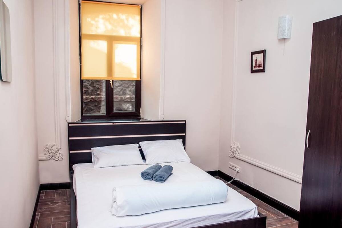A bedroom with a bed, a window with curtains, a picture on the wall, and a wardrobe.
