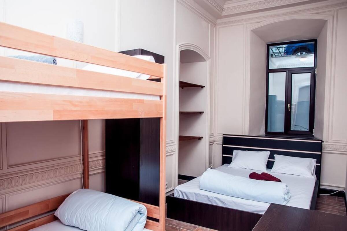 A hotel room with a bunk bed, a separate bed with white linens, a large window with a view, and built-in shelving.
