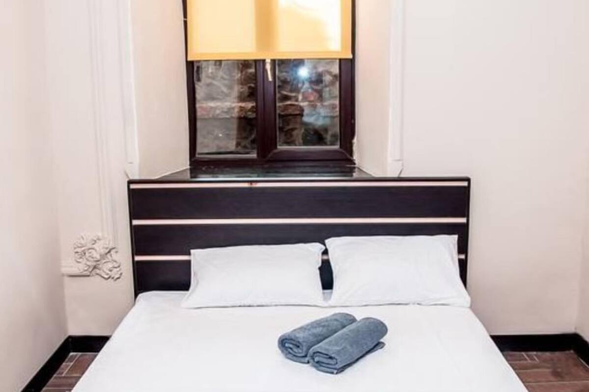 A neatly made bed with white linens and two rolled towels, a dark wooden headboard, and a window with a view of a building.