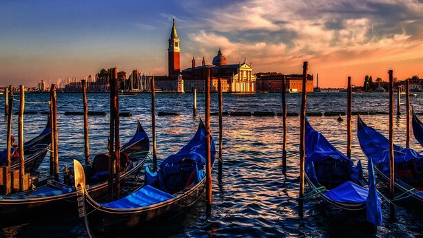 A waterfront scene with gondolas docked at a pier, a prominent tower, and a domed building in the background.