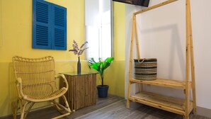 Una habitación con una silla de mimbre, una mesita con un jarrón de flores y un estante de madera con una cesta.