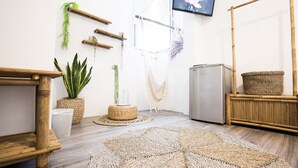 Una habitación con piso de madera, un tapete redondo tejido, una pequeña mesa, un refrigerador y varias plantas.