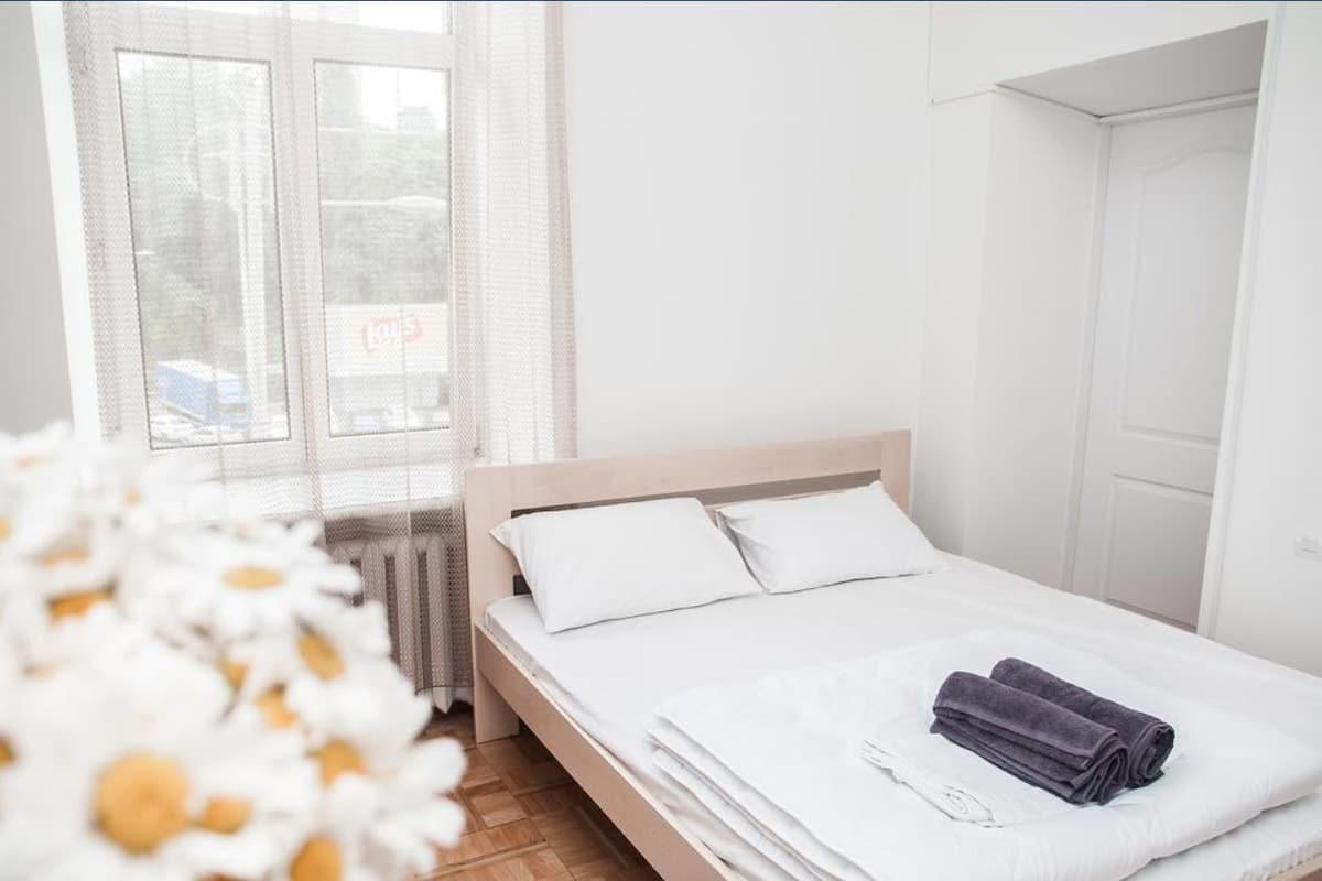 A clean, modern bedroom with a wooden bed, white bedding, and a window with sheer curtains.
