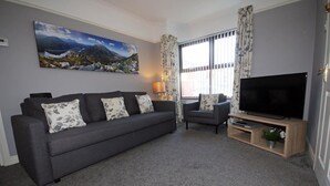 Una sala de estar con un sofá gris, un sillón más pequeño, un televisor de pantalla plana sobre un mueble de madera y un cuadro de un paisaje montañoso en la pared.