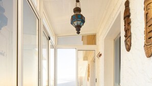 Een smalle gang met een betegelde vloer, een witte muur met een houten snijwerk, een hangende lantaarn en uitzicht door een raam.