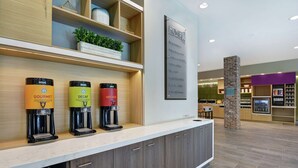Ein moderner Küchenbereich mit einer Kaffeestation, die drei Kaffeezubereiter mit den Aufschriften 'GOURMET', 'DECAF' und 'HEISSWASSER' zeigt. Es gibt eine Steinsäule und ein Schild an der Wand mit dem Schriftzug 'HOMER'.