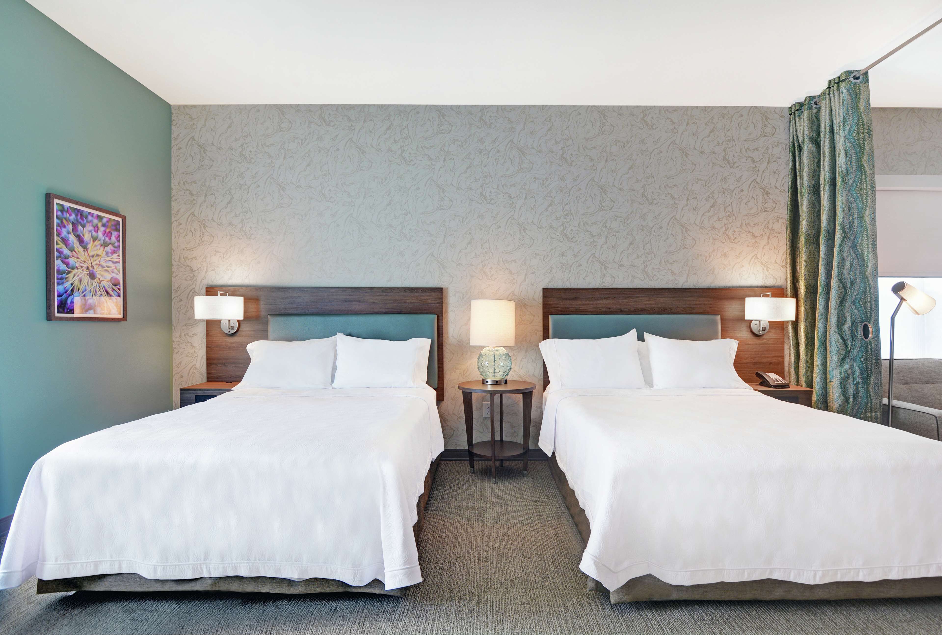 A hotel room with two beds, a small table with a lamp, and a painting on the wall.