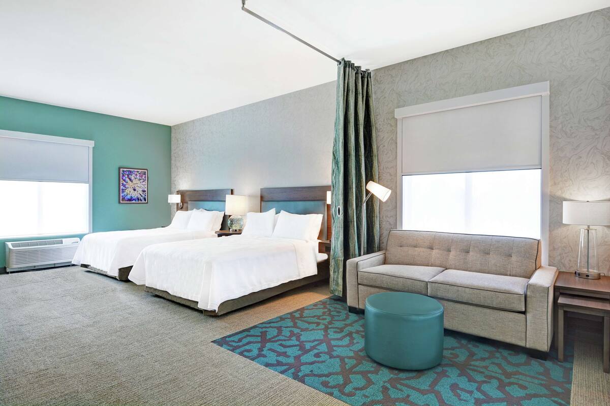 A hotel room with two beds, a sofa, a round ottoman, and a large window with curtains.