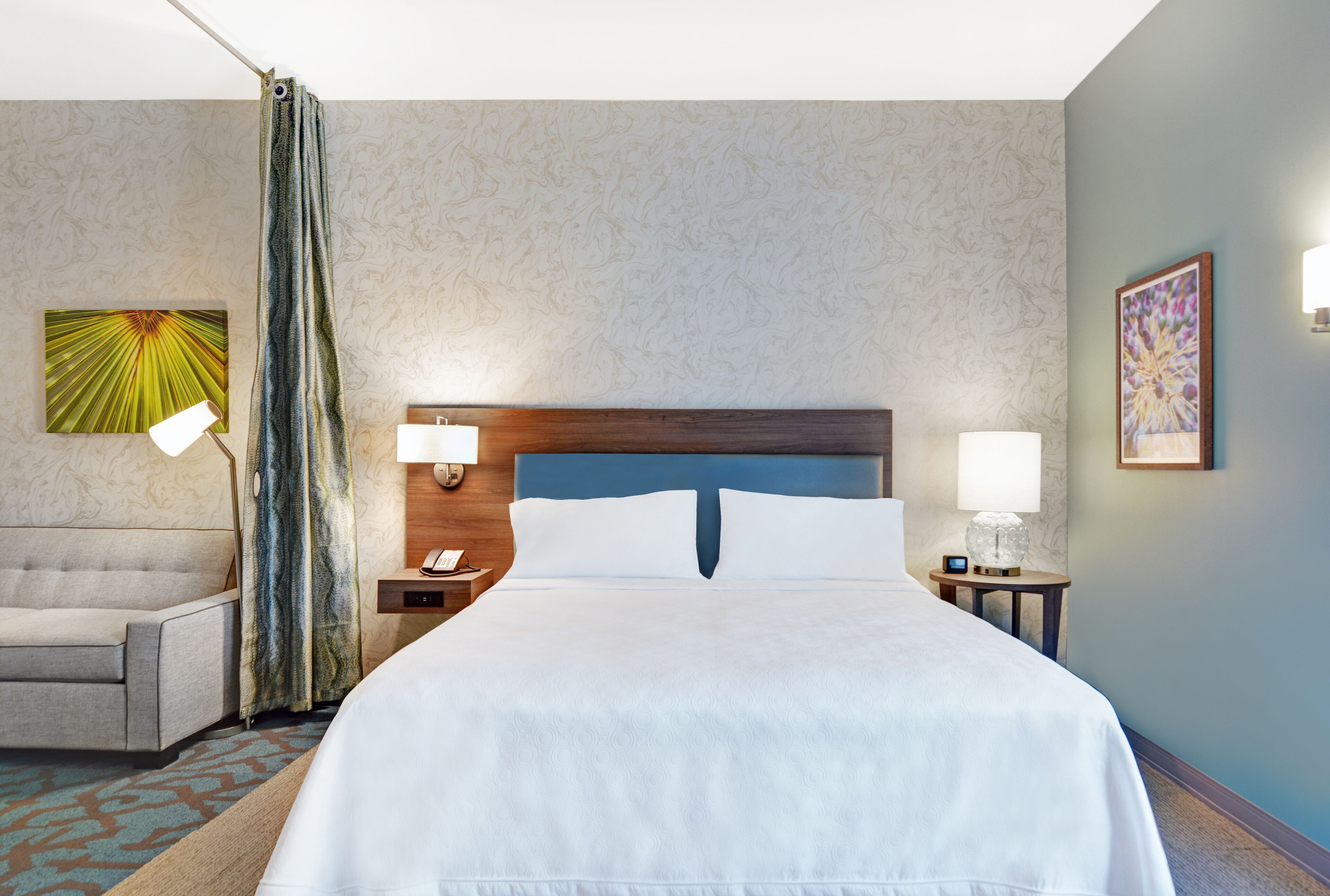 Une chambre d’hôtel avec un grand lit, une chaise, une table de chevet avec une lampe et un mur orné d’un tableau représentant des motifs floraux.