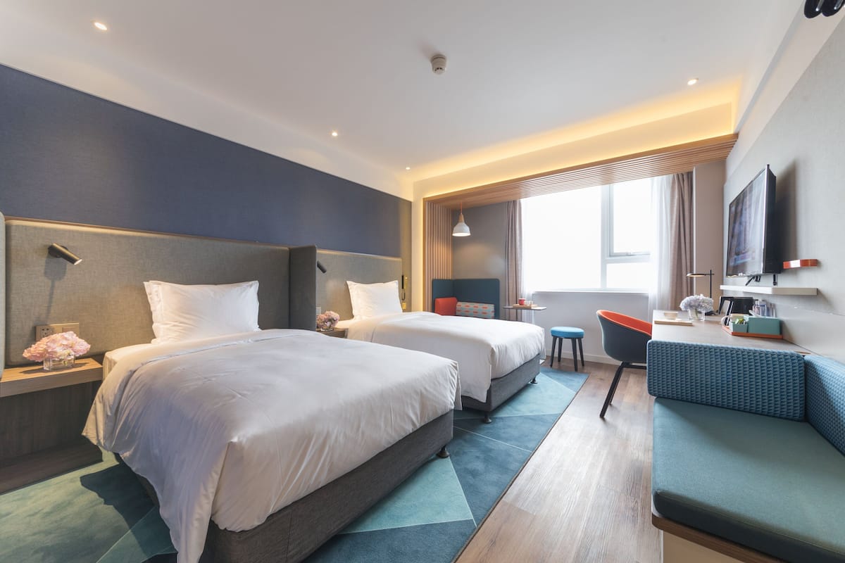 A modern hotel room with two beds, a sofa, a desk, and a TV.