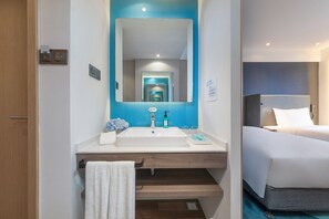 Bany d'hotel amb un mirall gran, un lavabo i un llit visible a través d'una porta oberta.