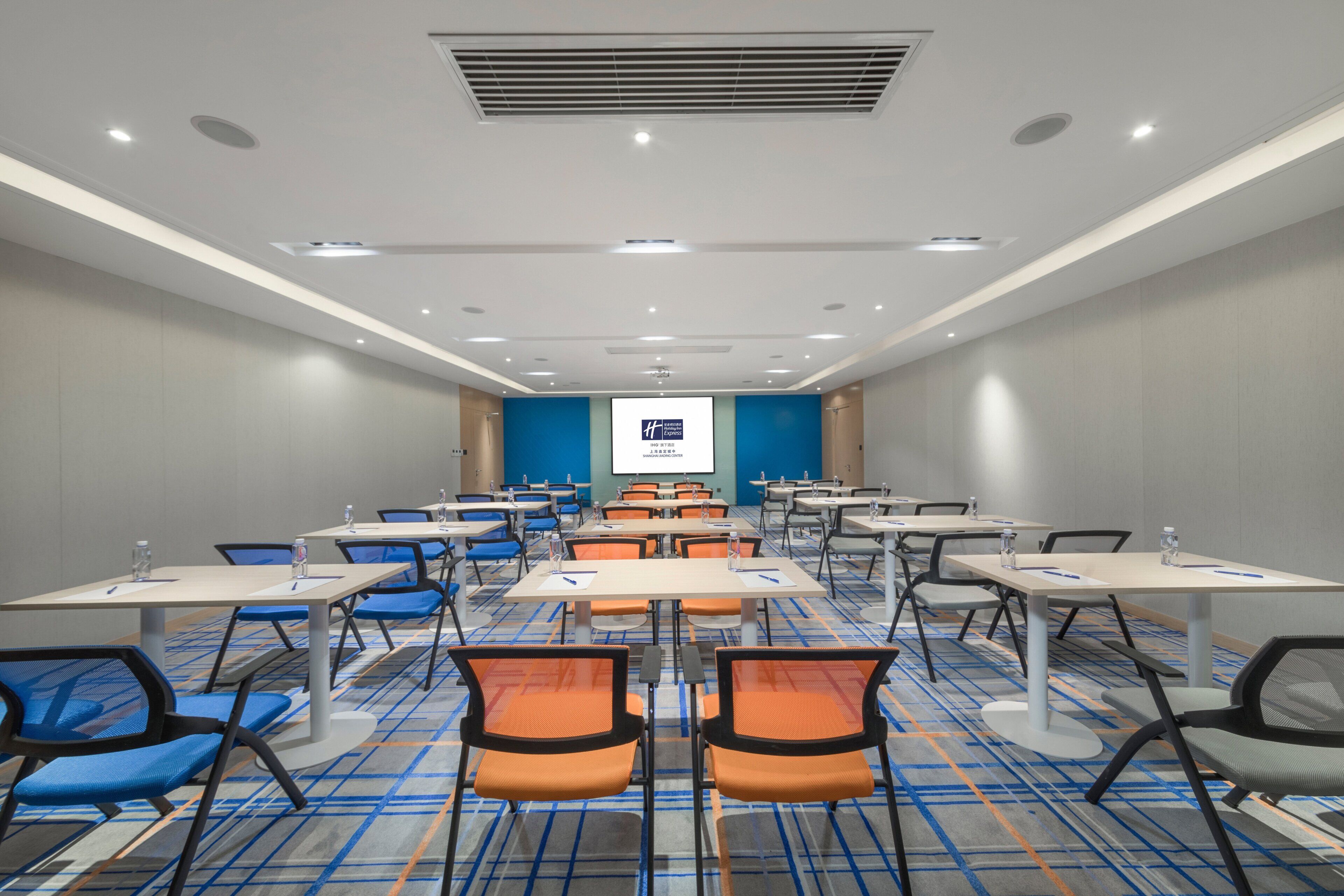 Een congreszaal met rijen tafels en stoelen, een scherm voor een beamer en een vloerbedekking.