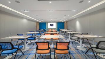 Een congreszaal met rijen tafels en stoelen, een scherm voor een beamer en een vloerbedekking.