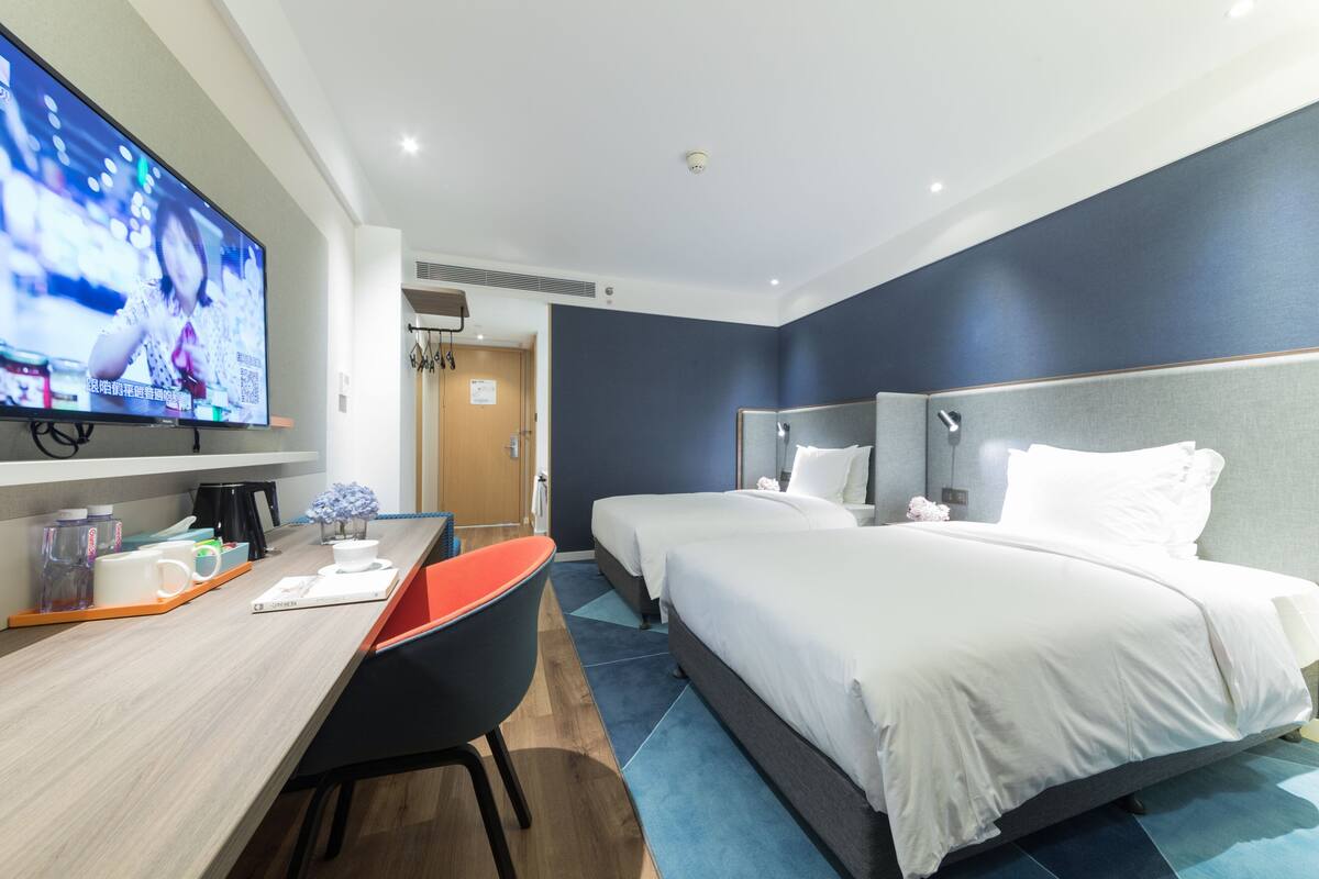 A hotel room with two beds, a desk with a chair, a flat-screen TV, and a coffee machine.