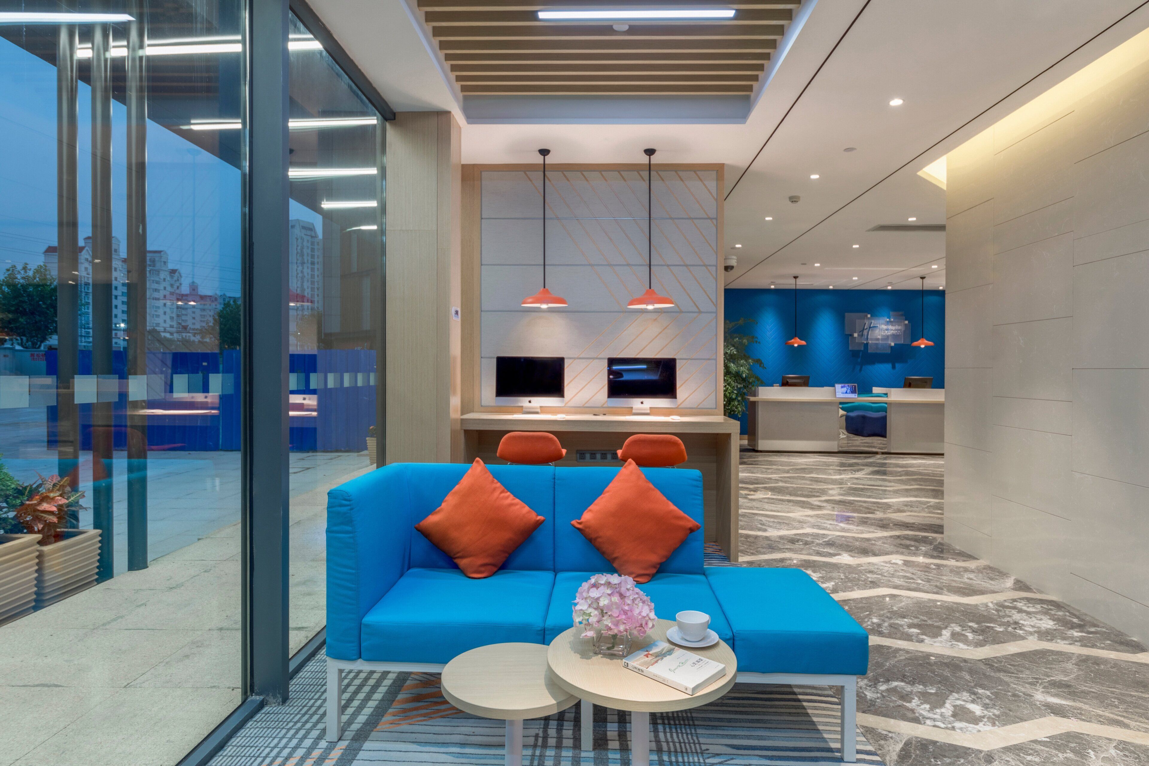 A modern lobby with a blue sofa, two orange pillows, a small round table with a book and a vase of flowers, and a reception desk with two monitors.
