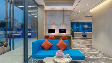 A modern lobby with a blue sofa, two orange pillows, a small round table with a book and a vase of flowers, and a reception desk with two monitors.