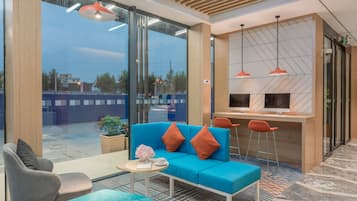 A modern hotel lobby with a blue sofa, grey armchairs, a small table with a vase, and a desk with computers.