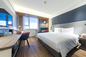 Habitació d'hotel moderna amb un llit gran, una taula de treball amb cadira, un televisor de pantalla plana i una finestra amb cortines.