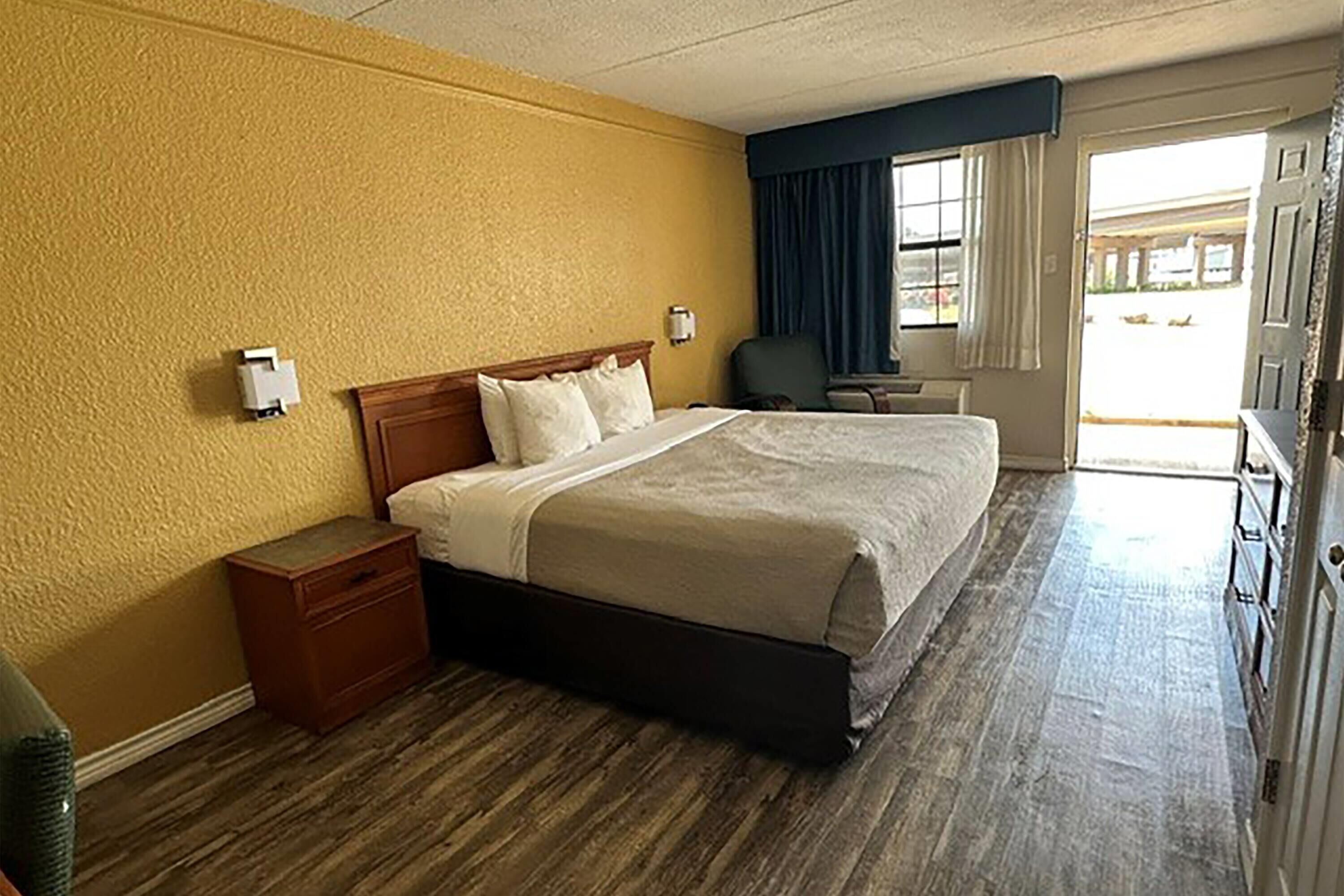 A hotel room with a large bed, wooden nightstand, and a view of the outside through a window with curtains.