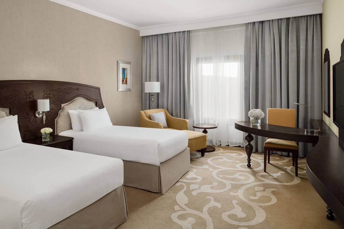 A hotel room with two beds, a desk, a chair, a lamp, and a TV.