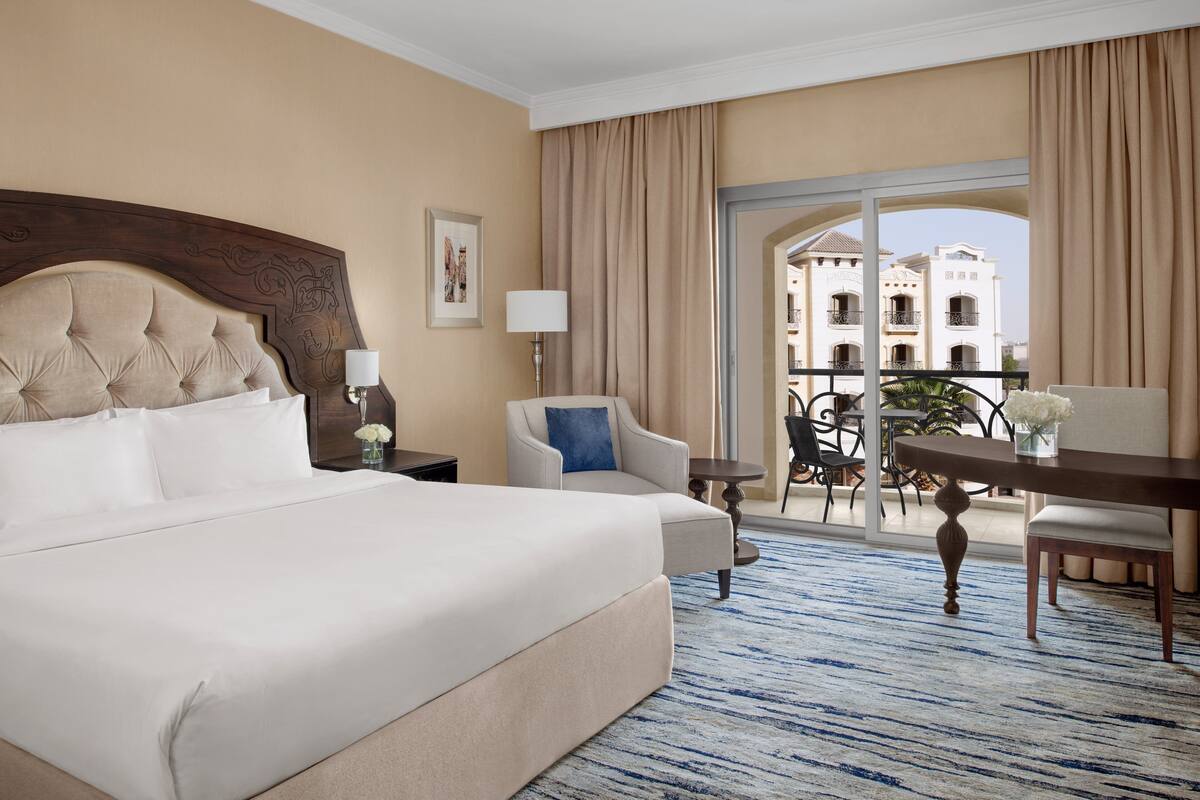 A hotel room with a large bed, a chair, a table, and a balcony with a view of a building.