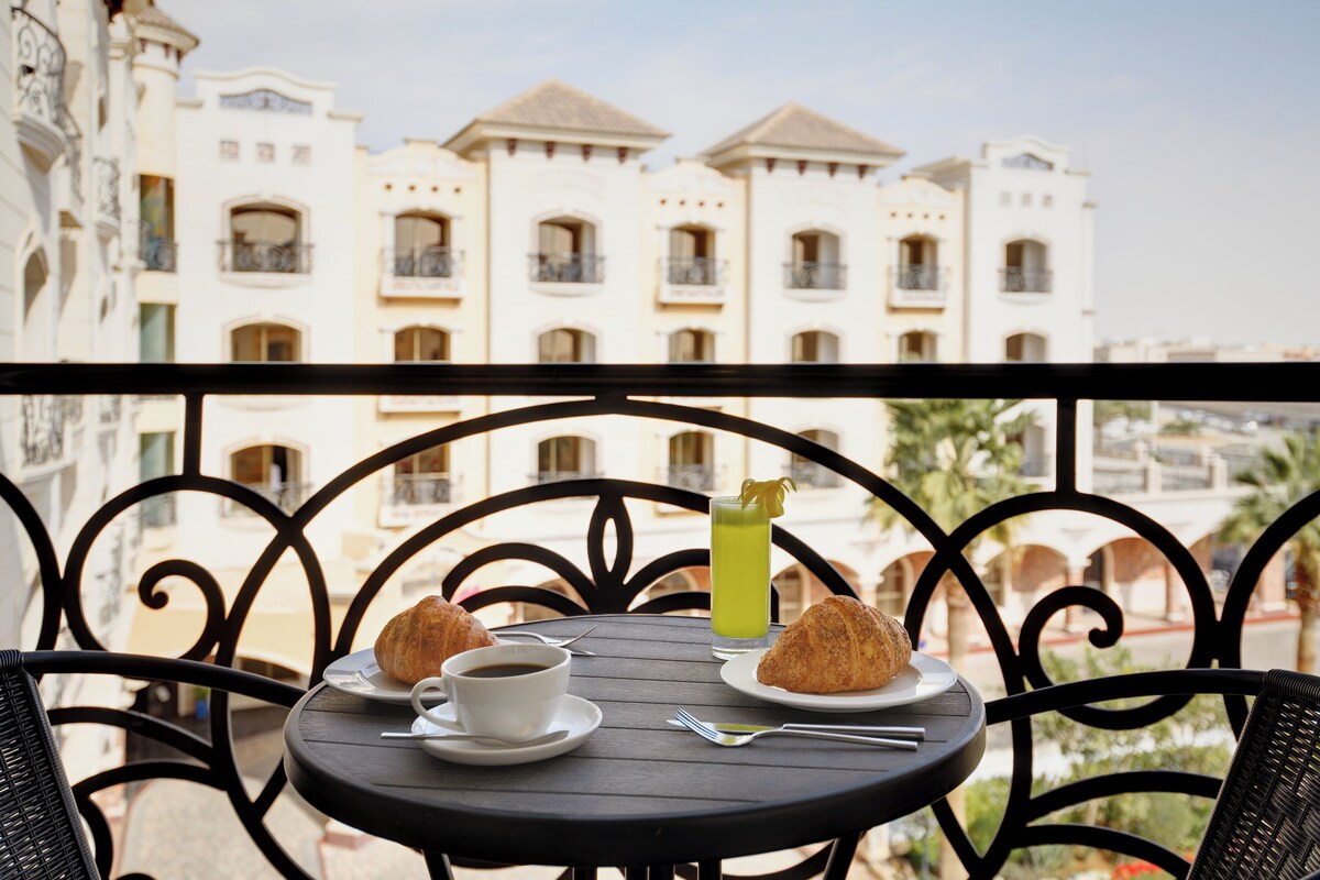 Een balkon met een tafel voor twee, met een croissant, een kop koffie en een glas groene groentesap.