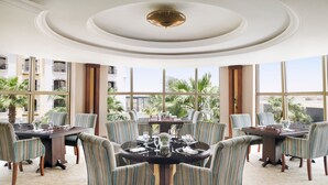 A dining area with round tables and striped chairs, large windows with a view of palm trees and buildings, and a decorative ceiling. - Crowne Plaza Riyadh Al Waha by IHG (Riyadh)