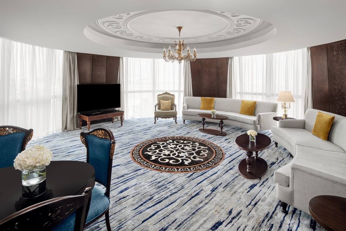 A spacious living room with a large TV, a sofa, a dining table with chairs, and a patterned carpet.