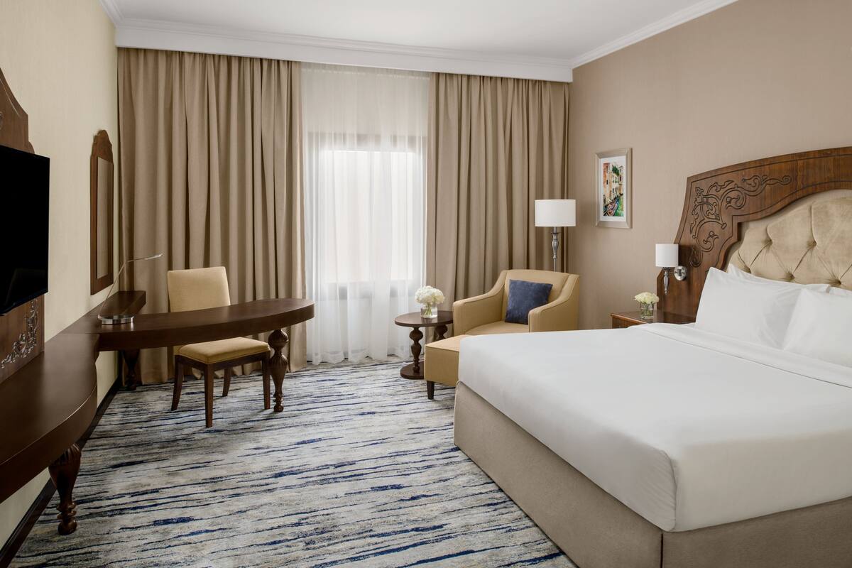 A hotel room with a large bed, a desk with a chair, a small table with a lamp, and a large window with curtains.