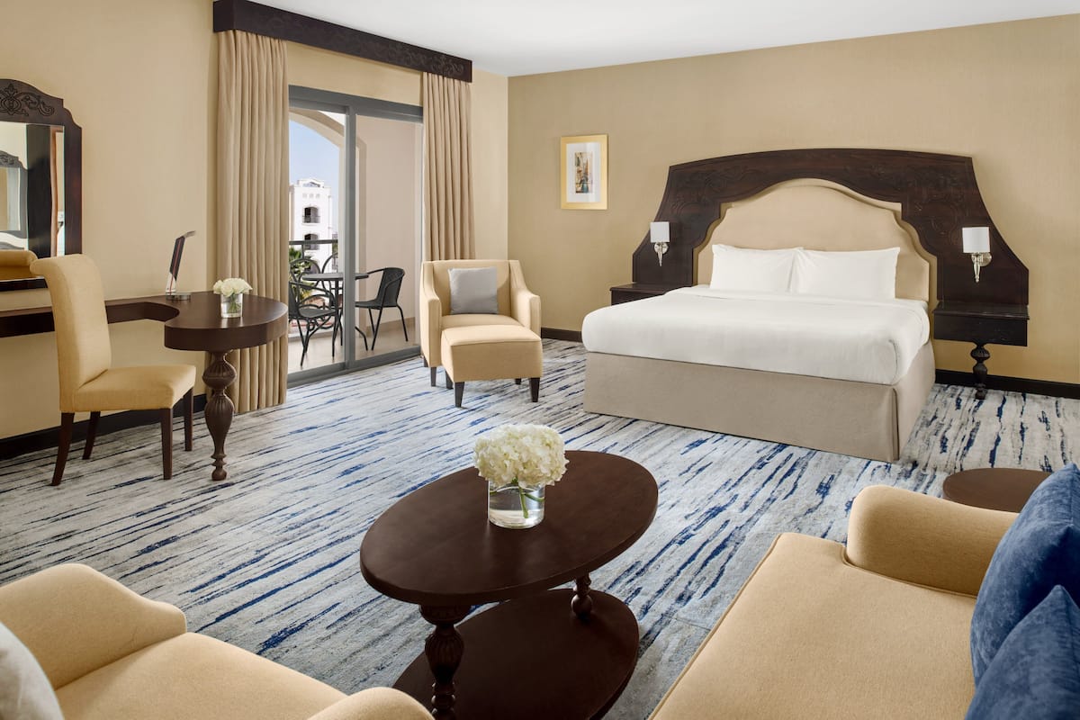 A hotel room with a large bed, two armchairs, a small table with a vase of flowers, and a view of the city through a large window.