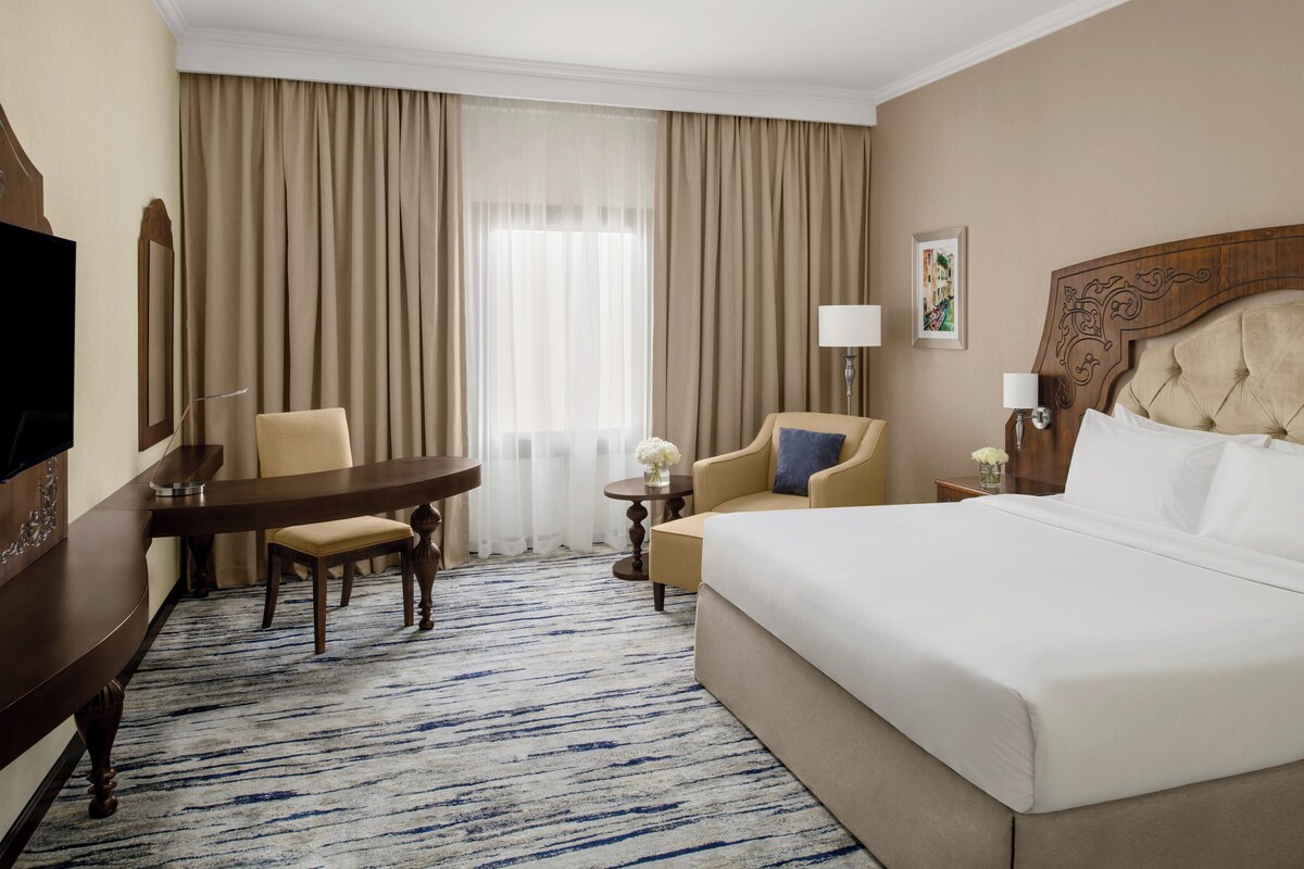 Een hotelkamer met een groot bed, een bureau met stoel, een kleine tafel met stoel, een lamp en een schilderij aan de muur.