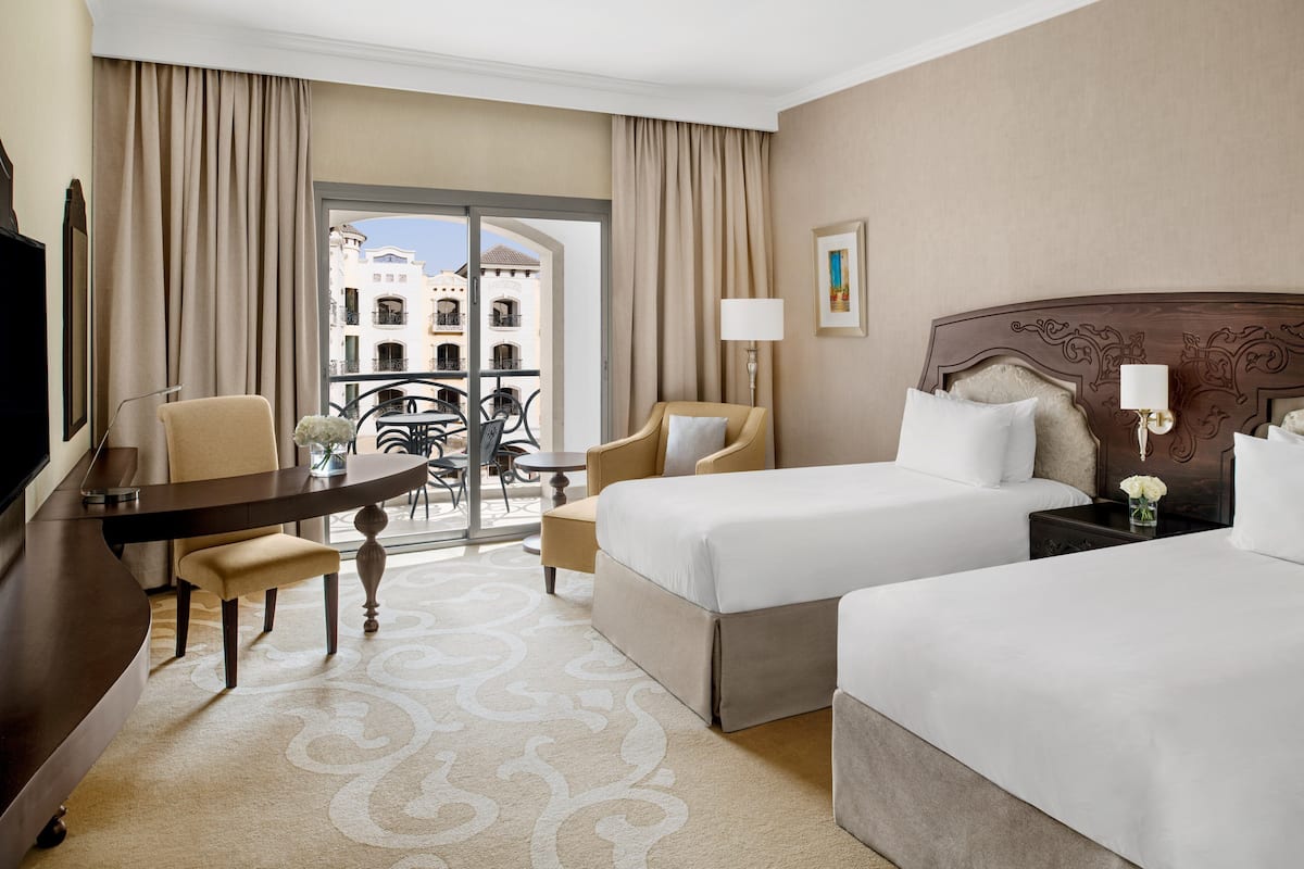 A hotel room with two beds, a dining table, chairs, a TV, and a balcony with a view of buildings.