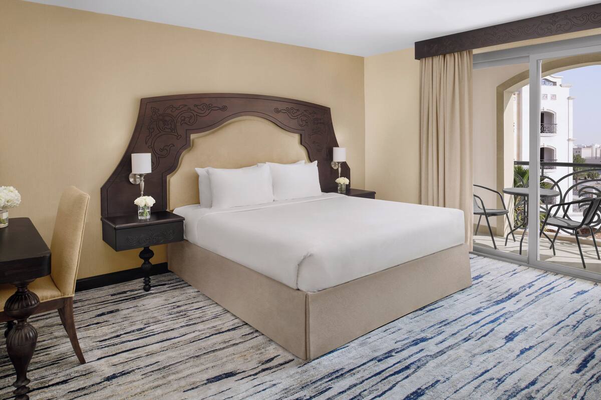 A hotel room with a large bed, a nightstand, a chair, and a balcony with a table and chairs.