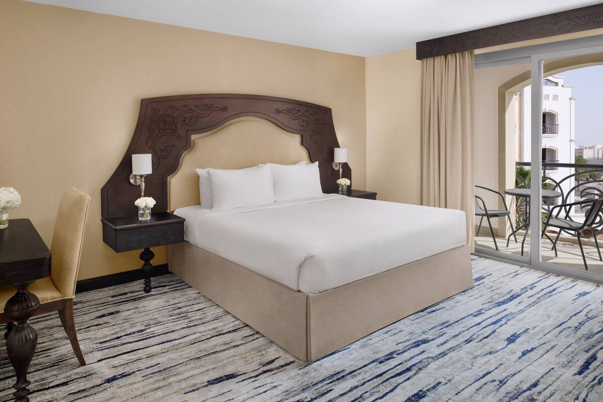 A hotel room with a large bed, a nightstand, a chair, and a balcony with a table and chairs.