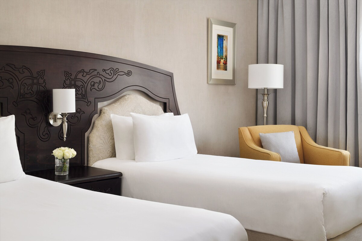 Een hotelkamer met twee bedden, een nachtkastje met een lamp, een stoel, een ingelijst schilderij aan de muur en een vaas met bloemen op het nachtkastje.