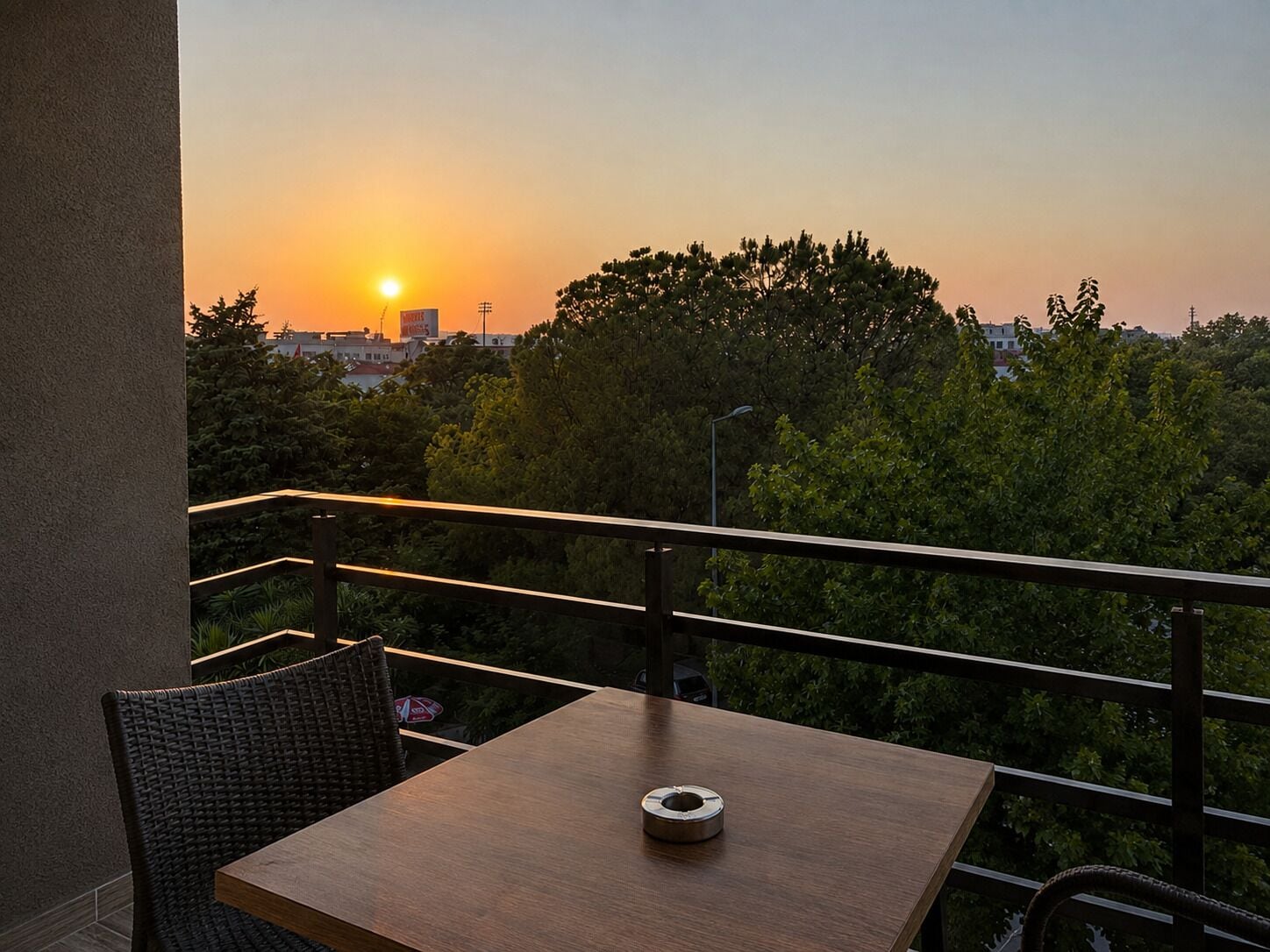 Un balcón con una mesa y una silla, con vista a un atardecer con árboles y edificios al fondo.