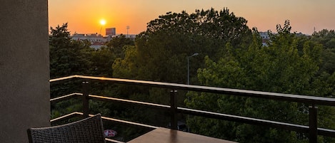 A balcony with a table and chair, overlooking a sunset with trees and buildings in the background.