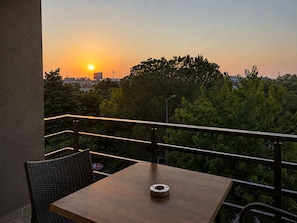 Ein Balkon mit Tisch und Stuhl, Blick auf einen Sonnenuntergang mit Bäumen und Gebäuden im Hintergrund.