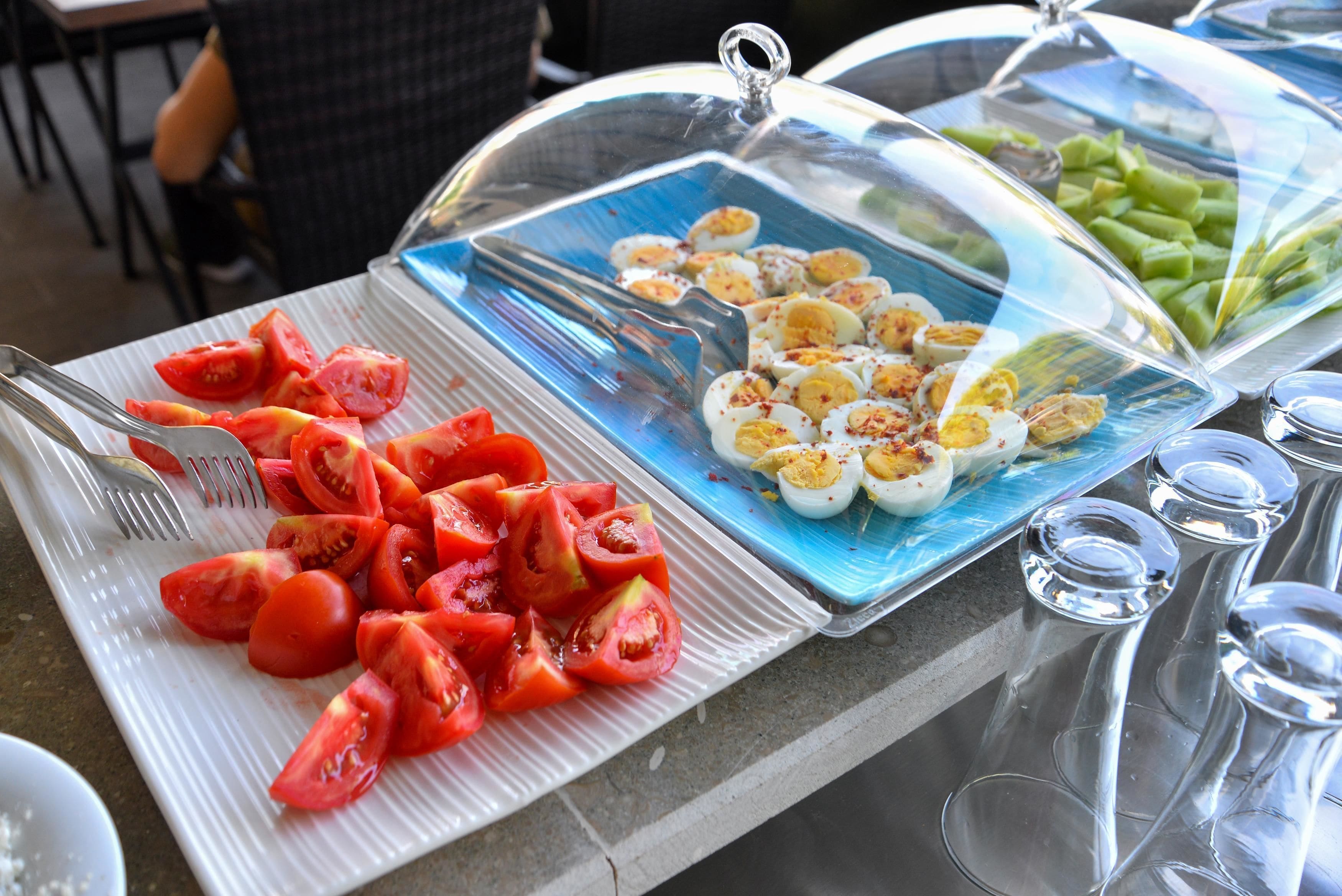 A buffet table with sliced tomatoes and hard-boiled eggs on a white plate, and a glass container with green vegetables.