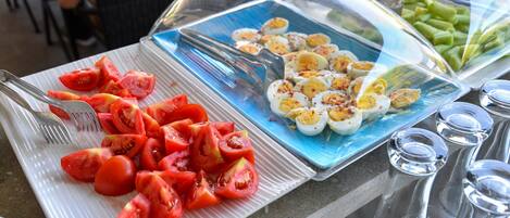 A buffet table with sliced tomatoes and hard-boiled eggs on a white plate, and a glass container with green vegetables.