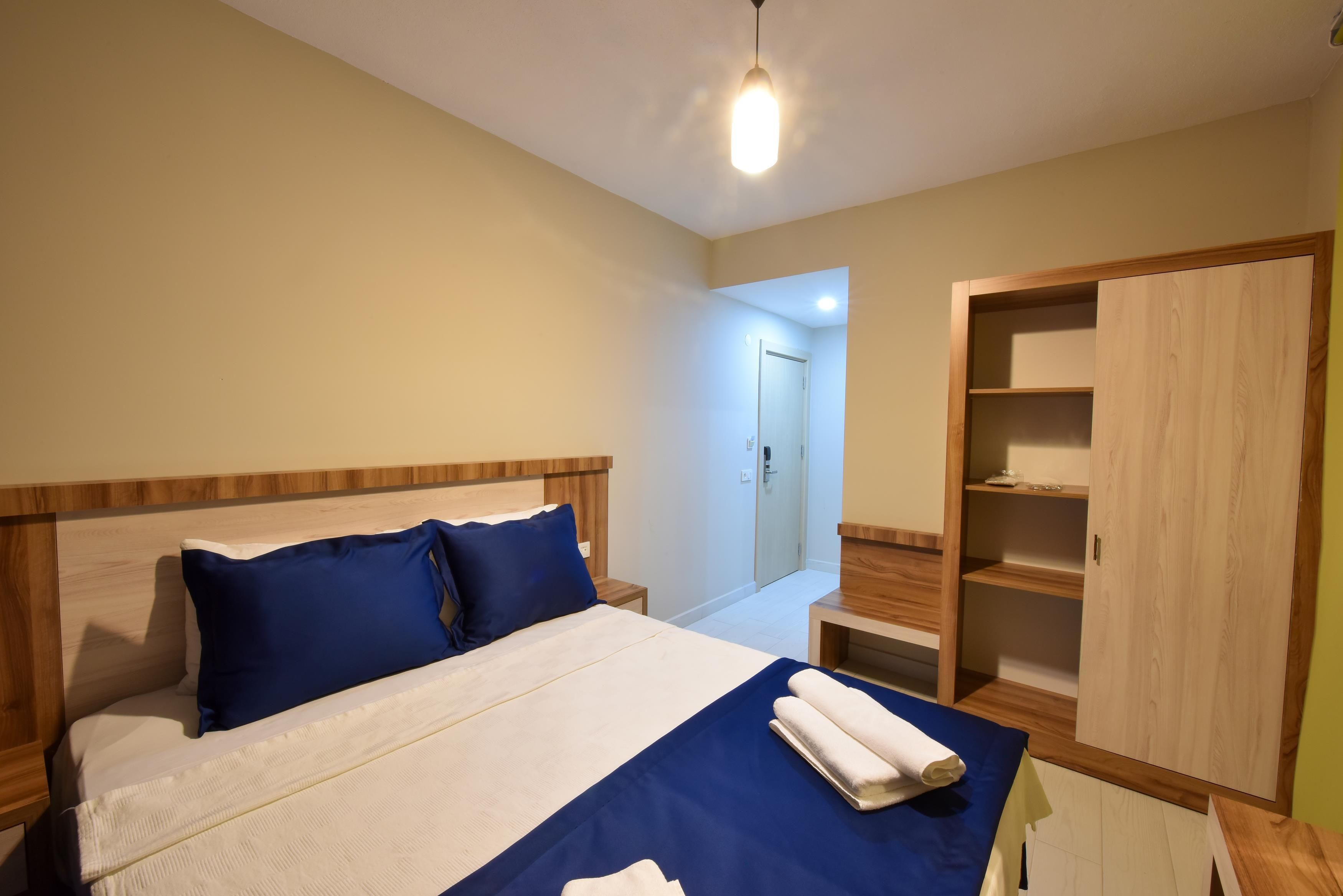 Ein ordentlich bezogenes Bett mit blauen Kissen, einem Holzkopfende und einer blauen Bettdecke. Dazu ein Holzschrank mit Einlegeböden und einer Tür auf der rechten Seite.