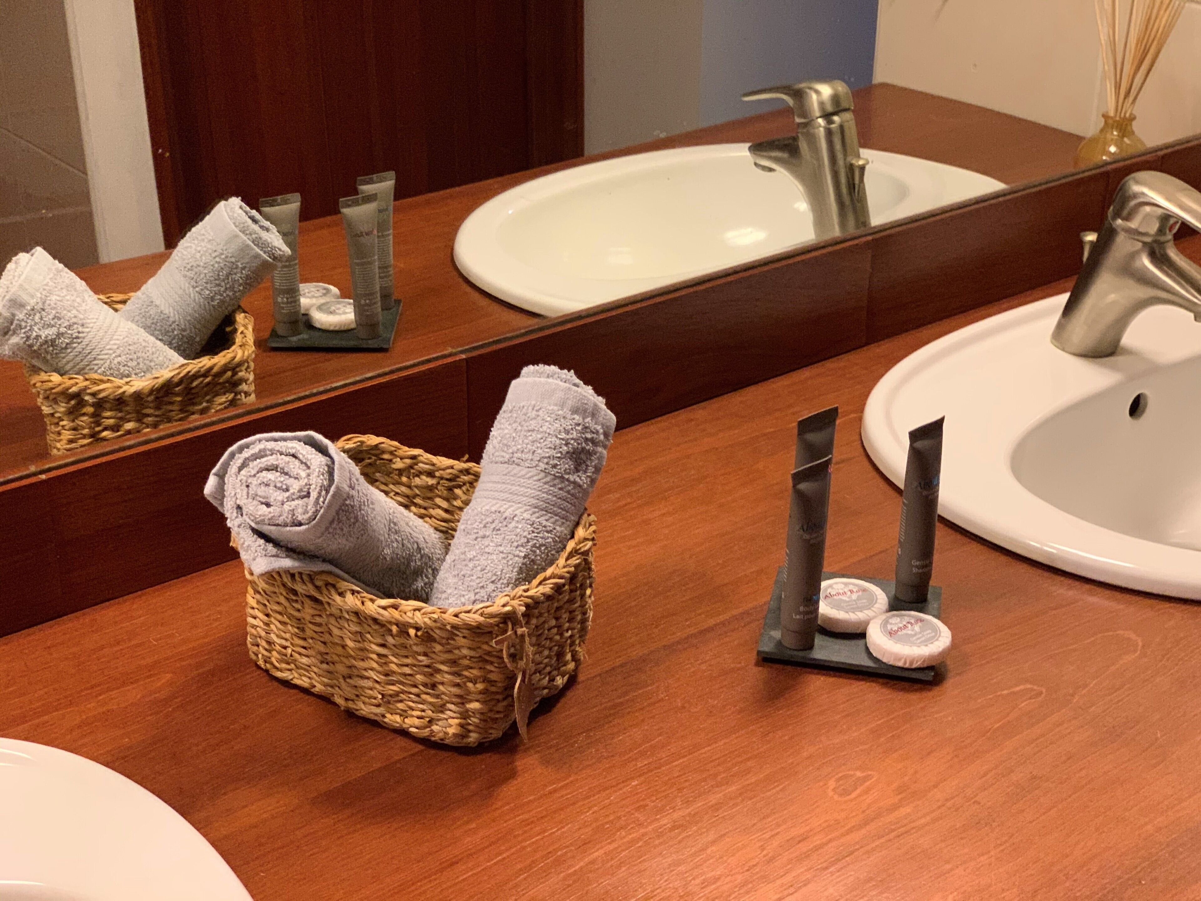 A bathroom with a wooden vanity, a white sink, a mirror, and a wicker basket holding rolled towels.