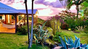 A cozy outdoor seating area with a hammock, surrounded by lush tropical plants and a view of the ocean at sunset. - Between luxury and authenticity.  Beautiful sea view property, 100m from the beach. (Deshaies)