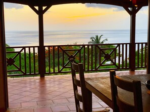 A covered outdoor seating area with a view of the ocean and palm trees.