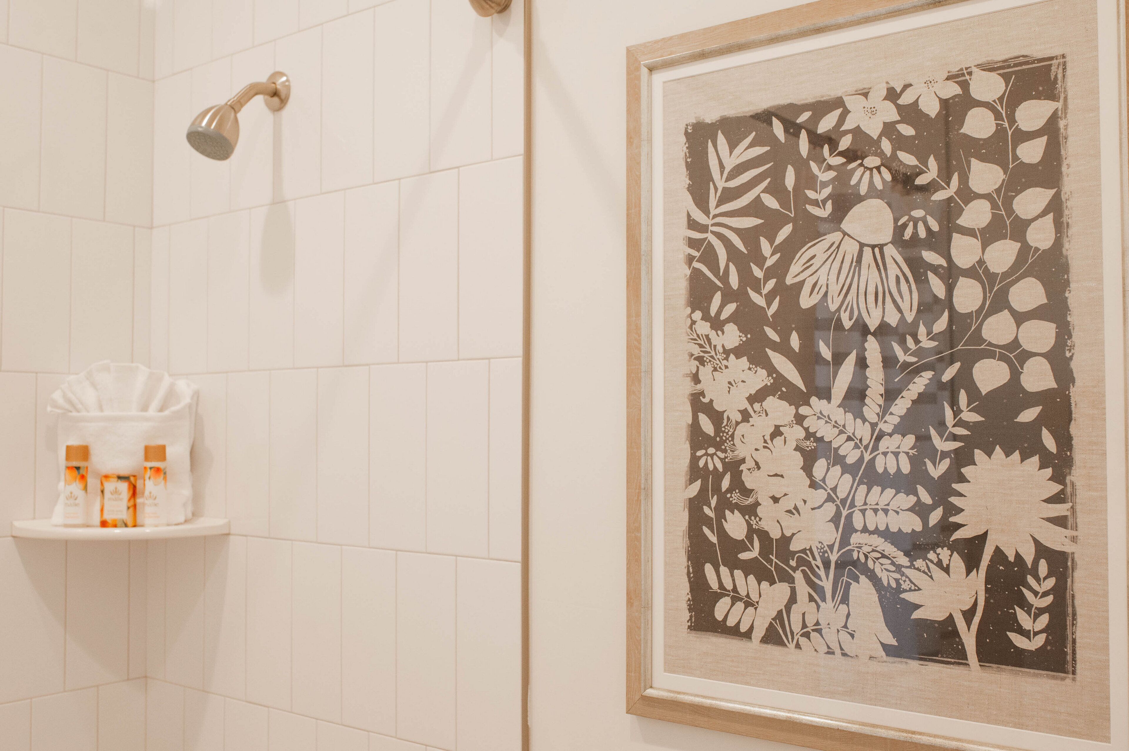 A bathroom with white tiles, a framed floral artwork, and a shelf with toiletries.